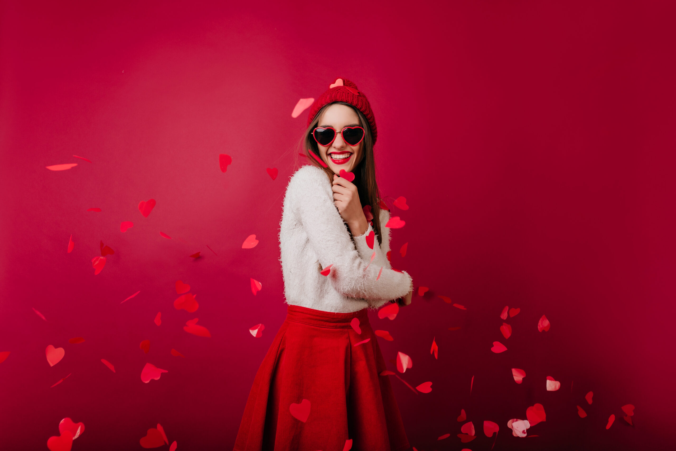 Emotional young woman in red hat and sunglasses standing on claret background at party. Studio shot of happy girl posing under heart confetti..